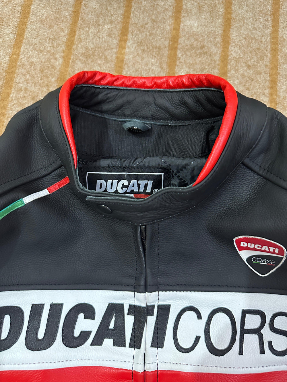Close-up of the Ducati Corse C5 Motorcycle Leather Jacket, highlighting the 'Ducati Corse' logo on the chest, the bold 'Ducati' branding, and the red and white accents. The image also shows the red-lined collar, emphasizing the jacket's premium quality and design details