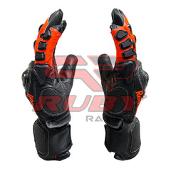 A close-up view of KTM Redbull motorcycle racing gloves in black and orange colors, focusing on the side profile. The gloves display the Redbull logo on the wrist, with detailed protective padding along the fingers and knuckles. The design highlights the gloves' ergonomic structure and reinforced areas for enhanced grip and safety while riding