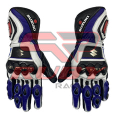 A pair of Suzuki motorcycle racing gloves in blue, white, and black colors. The gloves feature protective knuckle guards with the Suzuki logo, reinforced palms, and finger protection. They have a sporty design with a mix of leather and synthetic materials, Suzuki branding on the wrist strap, and the Suzuki "S" logo on the side. The gloves appear to be designed for high-performance riding and safety